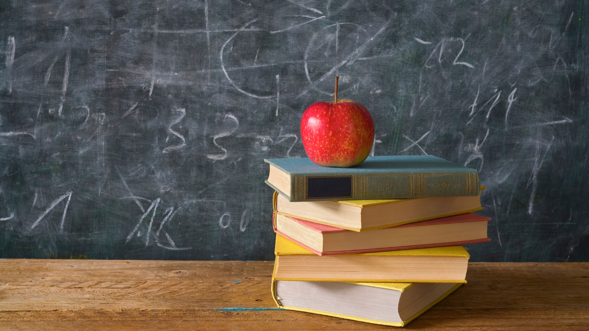 wood desk with black chalk board background; five text books stacked with a red apple on top;